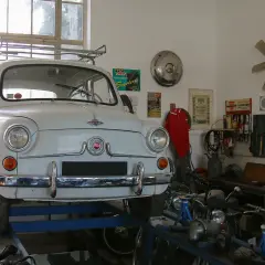 Biały Fiat 600 na podnośniku w warsztacie, gotowy do napraw. W tle narzędzia i motocykle, a nad nimi lampa zapewniająca oświetlenie kanału samochodowego.