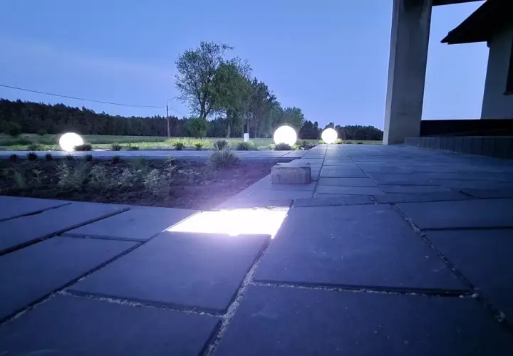 Nocny ogród rozświetlony kulistymi lampami. Ścieżka z kostki brukowej, na której widać wbudowane ledy, prowadzi do domu.