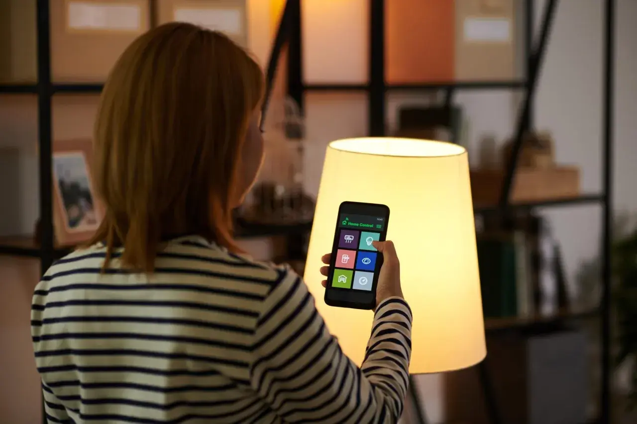 Kobieta steruje domem za pomocą smart life app na telefonie. Widok zza ramienia na ekran z ikonami.