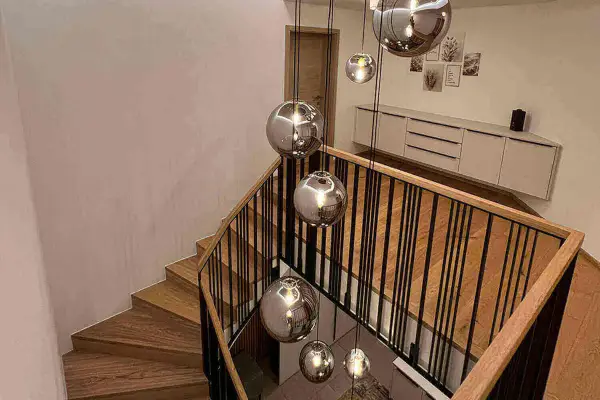 Nowoczesne lampy na klatkę schodową w kształcie kul, wiszące nad drewnianymi stopniami i czarną balustradą.